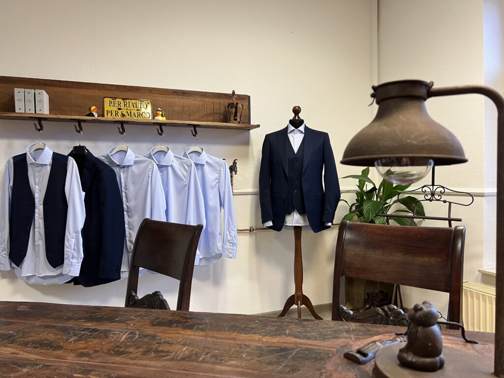 Verschiedene Hemden an einer Stange und daneben ein Maßgeschneiderter Anzug auf einem Ständer. Im Vordergrund ein alter Tisch mit zwei Stühlen und einer rustikalen Lampe